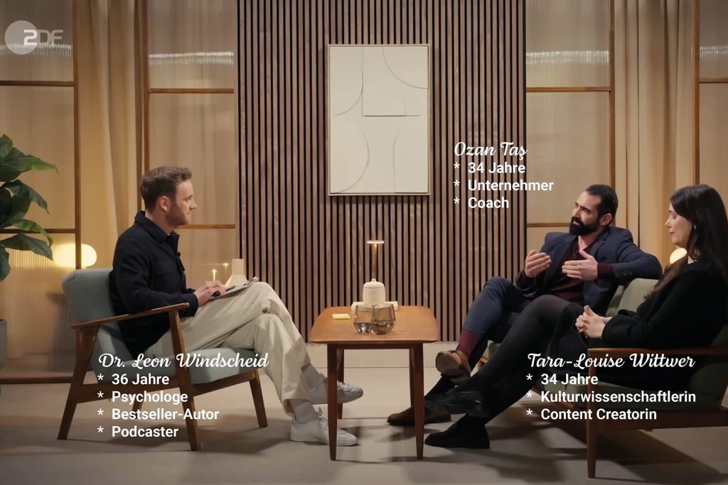 Zwei Männer und eine Frau sitzen sich gegenüber auf einem Sofa und einem Sessel. Sie diskutieren über die Frage "Fordern Frauen zu viel".