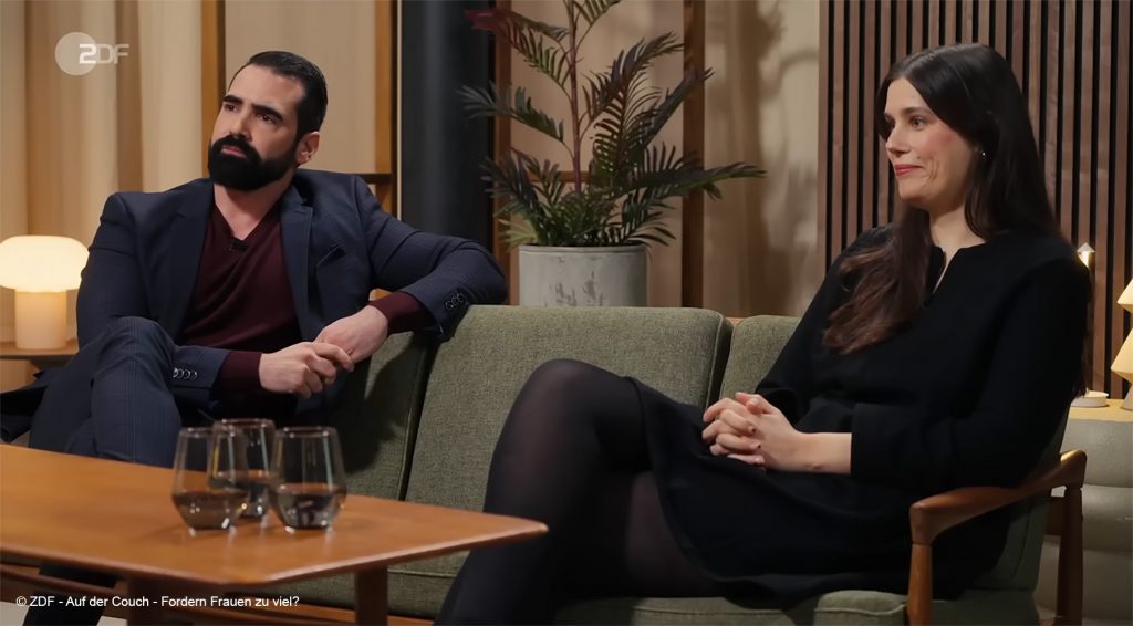 Ein Mann und eine Frau sitzen auf einem Sofa. Sie schauen auf den Moderator und diskutieren über die Frage "Fordern Frauen zu viel".