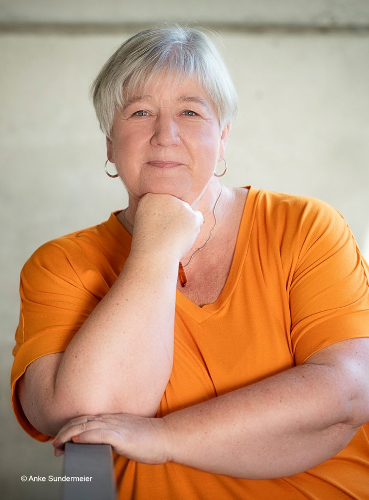 Eine Frau stützt ihren Kopf auf ihre Faust und lächelt in die Kamera. Sie trägt ein oranges T-Shirt.